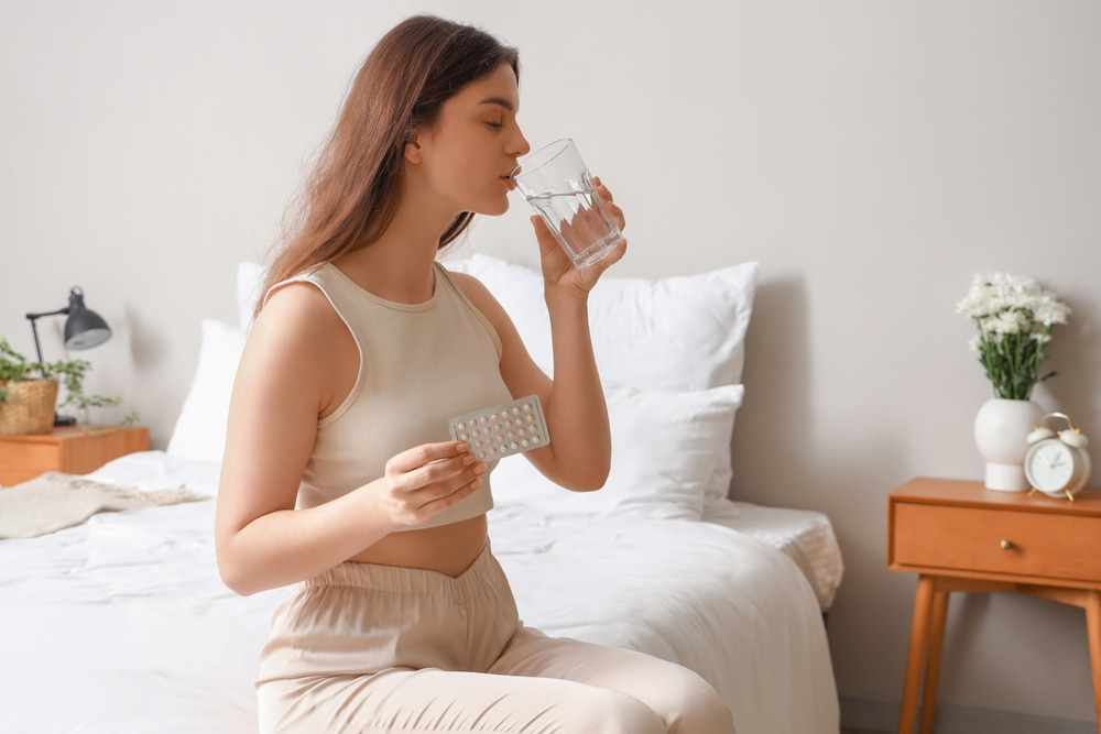 Mulher sentada na cama, tomando água, com uma cartela de remédio na mão.